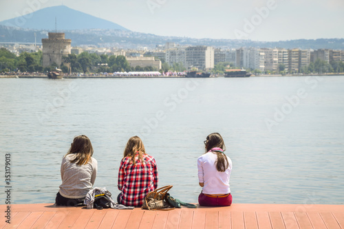 Three Teenagers Girls Friends Sitting On The Wooden Dock And Enjoying The City View In Front Of The Sea in Thessaloniki city in Greece, Europe.