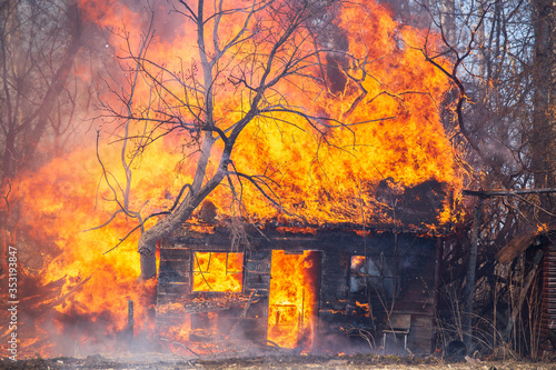 A small old house with square windows and trees in flames in a rural landscape