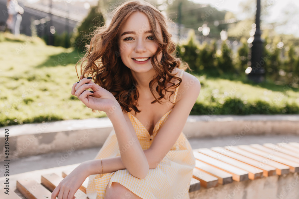 Naklejka premium Graceful ginger girl in yellow dress sitting in park. Lovely red-haired curly woman posing with inspired smile on nature background.