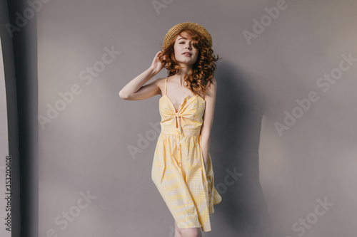 Shapely romantic lady in yellow dress having fun at photoshoot. Studio portrait of adorable ginger girl in hat isolated on gray background.
