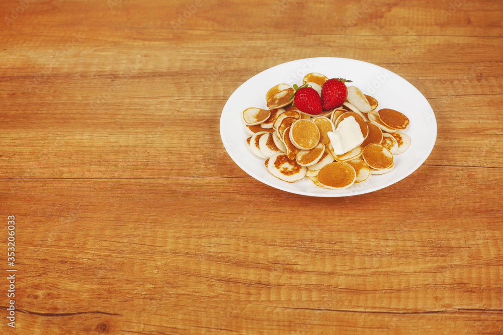 Delicious mini pancakes with strawberries and butter are placed on a white plate on a wooden background with space for text. Breakfast for children, trendy Breakfast, trendy food