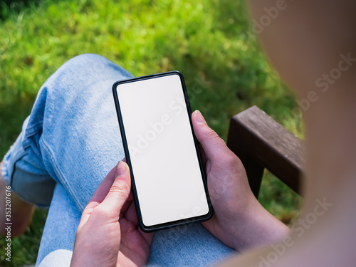 Young woman sitting outside in a garden on her phone with a blank white screen for mockup app and website designs. Concept showing technology being used in nature.