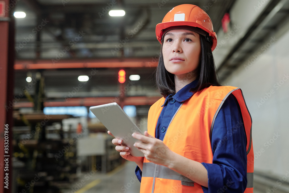 Pensive Asian female process engineer in work helmet and safety vest using digital tablet at factory Pensive Asian female process engineer in work helmet and safety vest using digital tablet at factory