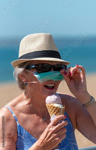 Southsea, Portsmouth, Southern England, UK. May 2020. Woman eating ice cream whilst wearing a mask and rubber protective gloves during the Corvid-19 outbreak. On the beach in Southsea. photo