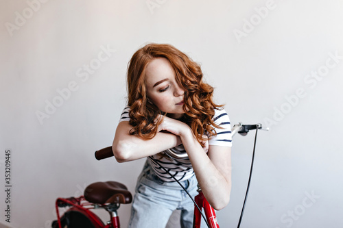 Good-looking ginger woman posing with bicycle on white background. Indoor photo of wonderful lady standing in studio with bike.