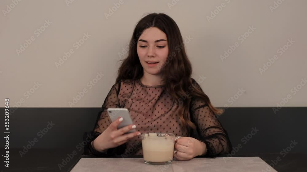 A charming young girl drinks coffee in a glass cup sitting at a table in a cafe and calls on the phone smiling. Close-up A charming young girl drinks coffee in a glass cup sitting at a table in a cafe and calls on the phone smiling. Close-up