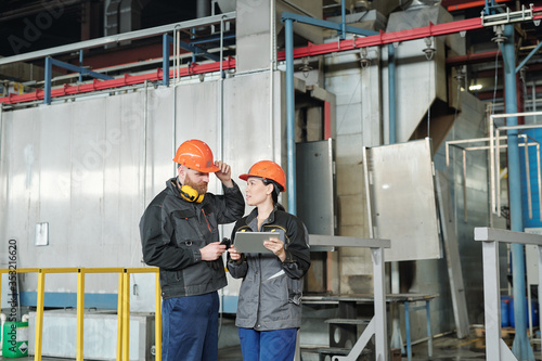 Asian woman in hardhat using digital tablet and discussing technical task with coworker in modern workshop