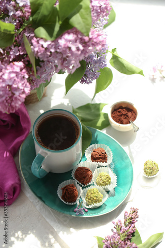 On a white background is a cup with a coffee, on a turquoise plate of candy and truffle, and in a vase is a lilac. 