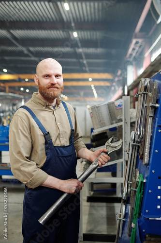Portrait of bald bearded man holding big metal wrench and standing in industrial shop