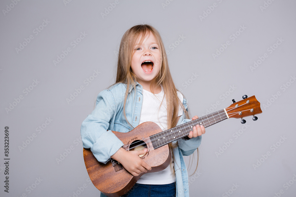 Fototapeta premium The child plays on the ukulele and sings songs. Joyful girl stands in a blue shirt on a gray background
