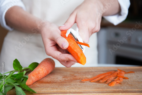 Hands of a young woman in an apron and on the background of the kitchen, peeling vegetables using a food peeler. Cleans the peel from carrots. Cooking fresh carrots before serving. photo