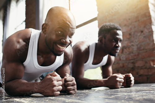Two African-Americans in white T-shirts train the abs and back muscles while standing with their forearms and legs on the floor.