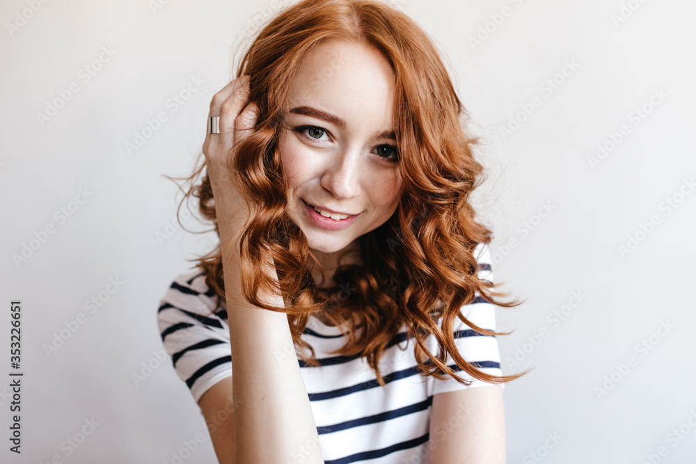 Fototapeta premium Wonderful dark-eyed girl touching her ginger hair with gently smile. Indoor photo of positive european lady in striped t-shirt.