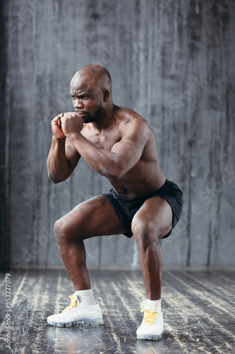 Strong and muscular dark-skinned man with a naked torso does squats while holding hands in his face in white sneakers