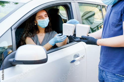 Young attractive girl in car gets 2 wok in box udon noodles with tempuru, shrimps, in hands and smiles. Courier in medical gloves. Udon noodles in white box delivery. Wok box udon advertise 1+1. © Rabizo Anatolii