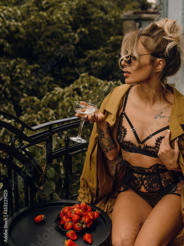 Young beautiful girl in black lingerie and a beige trench coat drinks champagne on a balcony in the morning