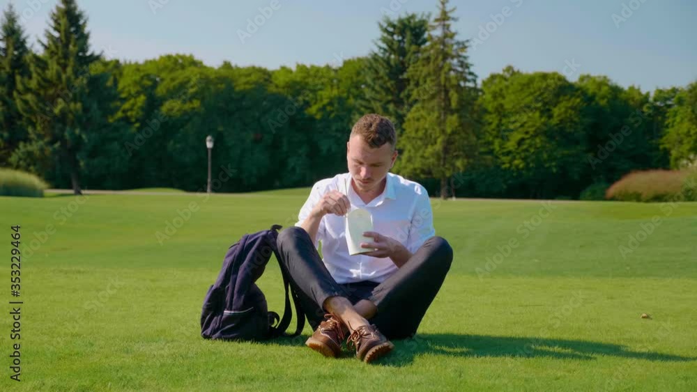 Businessman is eating vegetarian lunch with eco-friendly packaging. Freelancer sits in park on green grass and eats salad with tomatoes, cucumbers and lettuce in paper packaging with wooden sticks Businessman is eating vegetarian lunch with eco-friendly packaging. Freelancer sits in park on green grass and eats salad with tomatoes, cucumbers and lettuce in paper packaging with wooden sticks