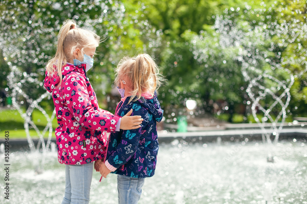 Two little girls in medical masks walk in the park near the fountain and hug each other Two little girls in medical masks walk in the park near the fountain and hug each other
