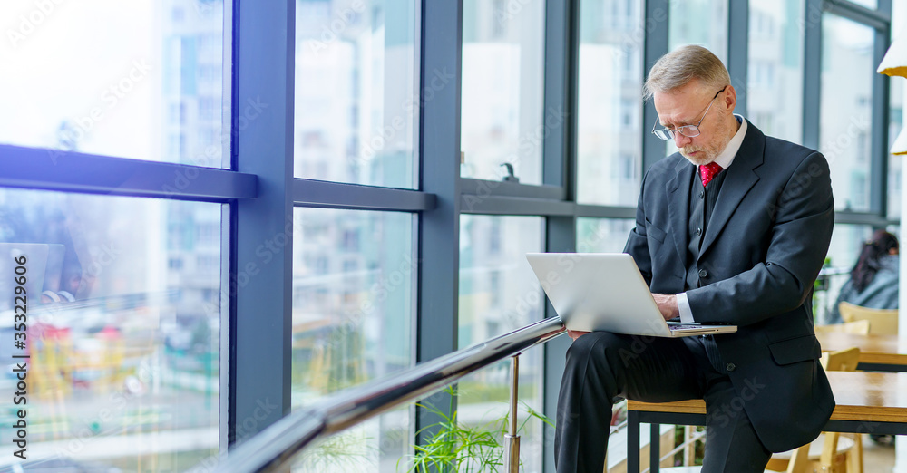 Thoughtful middle aged businessman in suit. Sitting near the window in office working on laptop. Panoramic city view background. Business photo.