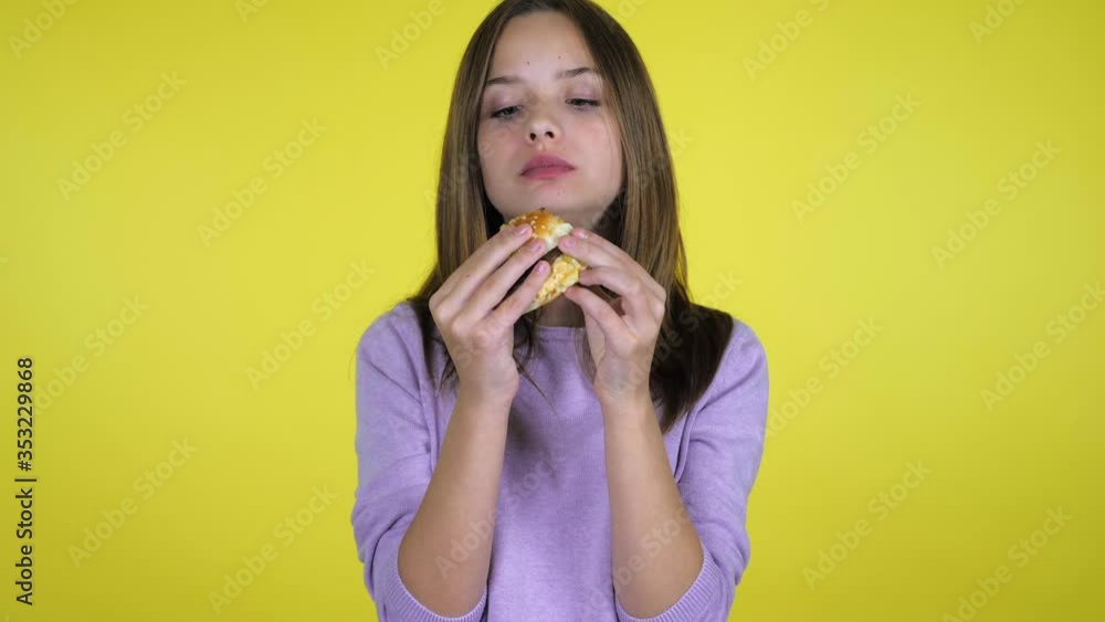 Teen girl in a pink sweater bites and eats burger on yellow background with copy space. Place for text or product. 4K slow motion footage Teen girl in a pink sweater bites and eats burger on yellow background with copy space. Place for text or product. 4K slow motion footage