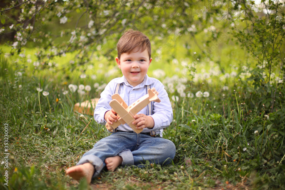 Little cute blond boy playing with a wooden plane in the summer park on the grass on a sunny day, focus on the child, blurred background Little cute blond boy playing with a wooden plane in the summer park on the grass on a sunny day, focus on the child, blurred background
