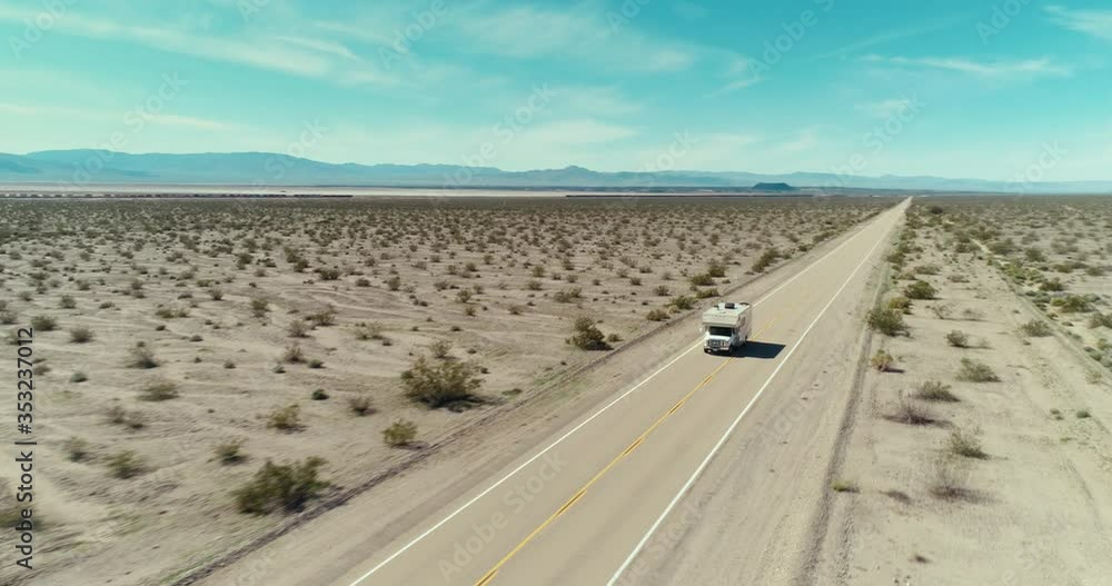 AERIAL - A drone tracks a small brown camper from the front in the middle of the Mojave desert on a long road on a clear and blue day in the summer with mountains in the back AERIAL - A drone tracks a small brown camper from the front in the middle of the Mojave desert on a long road on a clear and blue day in the summer with mountains in the back