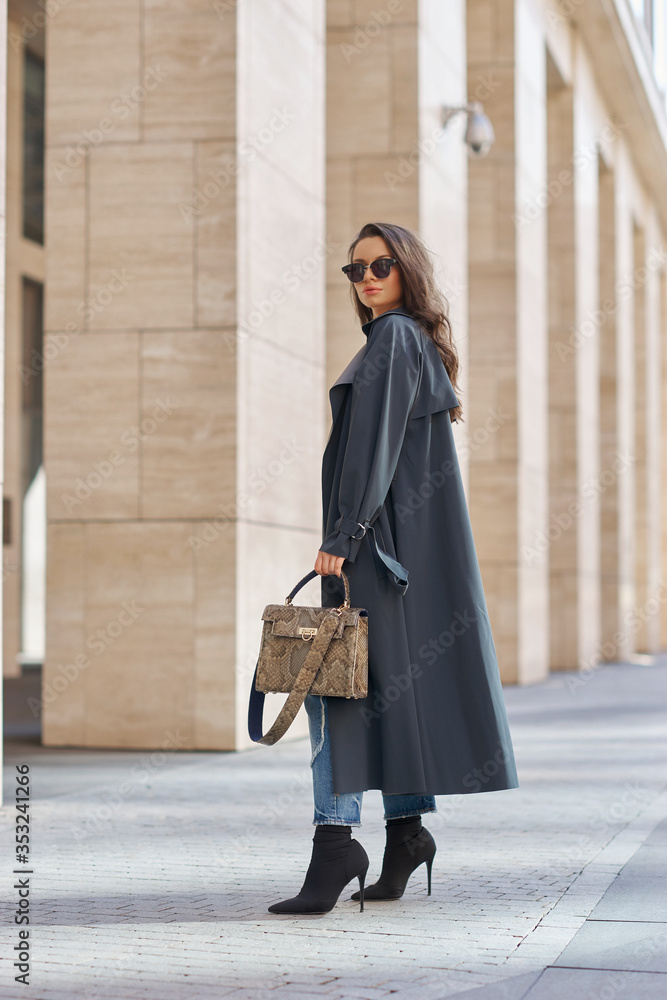 Naklejka premium Full length portrait. Woman walking city street. Female model holding leather reptile handbag. Stylish girl in gray raincoat and sunglasses. Caucasian brunette lady with wavy hair