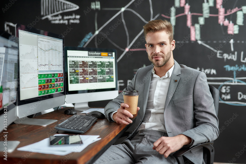 Solving money out. Portrait of successful young trader looking at camera and drinking coffee while sitting in front of multiple monitors in the office. Blackboard full of charts in background. Solving money out. Portrait of successful young trader looking at camera and drinking coffee while sitting in front of multiple monitors in the office. Blackboard full of charts in background.