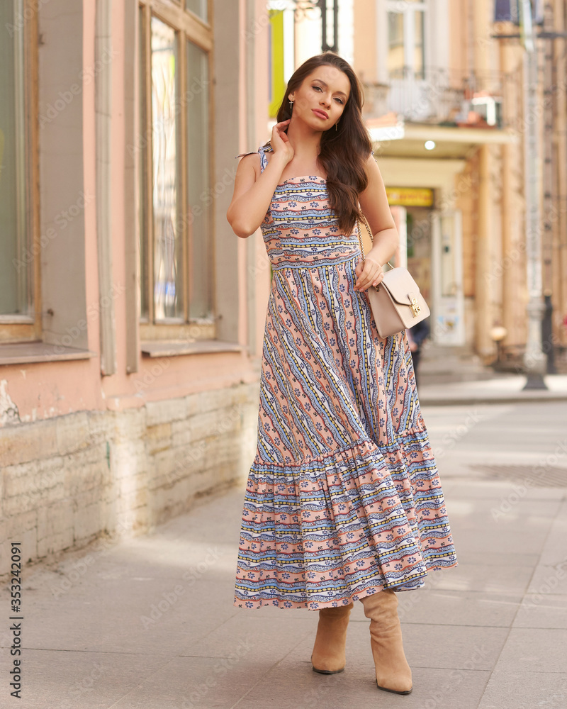 Elegant tender woman with makeup and long wavy hair walking city street. Pretty girl wearing blue and pink pastel colors sundress. Soft light portrait. Street look. Fashionable female model