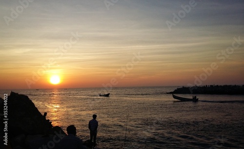 A silhouette landscape image of people watching sunset on a beach with fishing boats in Kerala, India.