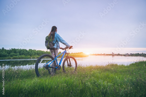 Woman with bicycle on the hill near the river at sunset in spring. Landscape with sporty girl with backpack riding a mountain bike, dirt road, green grass, water in summer. Sport and travel. Cycle