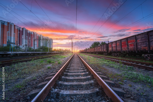 Railway station with freight trains and multicolored buildings at sunset. Railroad in summer. Heavy industry. Landscape with train, railway platform, sky with colorful clouds at dusk. Transportation