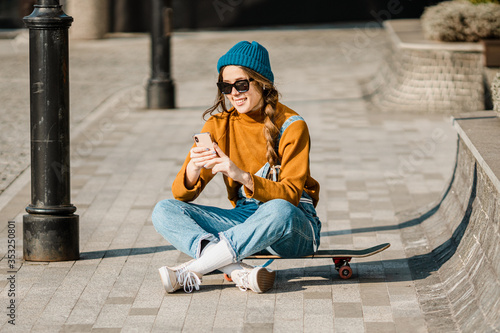 Girl sitting on skateboard and use mobile phone. Outdoors, urban lifestyle. cute skater girl sitting on skate board checking smart phone listening to music using internet and takes a photo