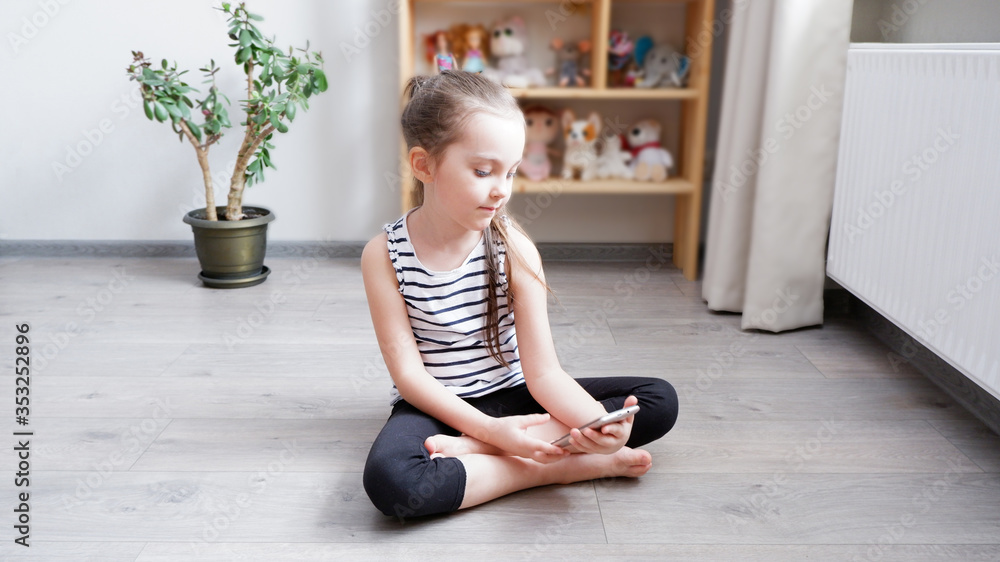 Distance learning. Little girl sitting on a wooden floor with a phone, watching a cartoon or making a video call on a computer Distance learning. Little girl sitting on a wooden floor with a phone, watching a cartoon or making a video call on a computer