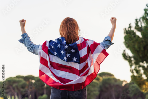 Young happy woman from behind with the United States flag raising arms excited in nature. 4th of july independence day of the united states