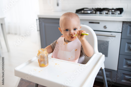 baby girl sits in the kitchen in a feeding chair and eats photo