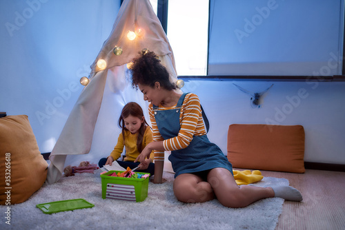 Excellent babysitter you can count on. African american woman baby sitter entertaining caucasian cute little girl. Kid reading a book with her nanny sitting in wigwam, tent