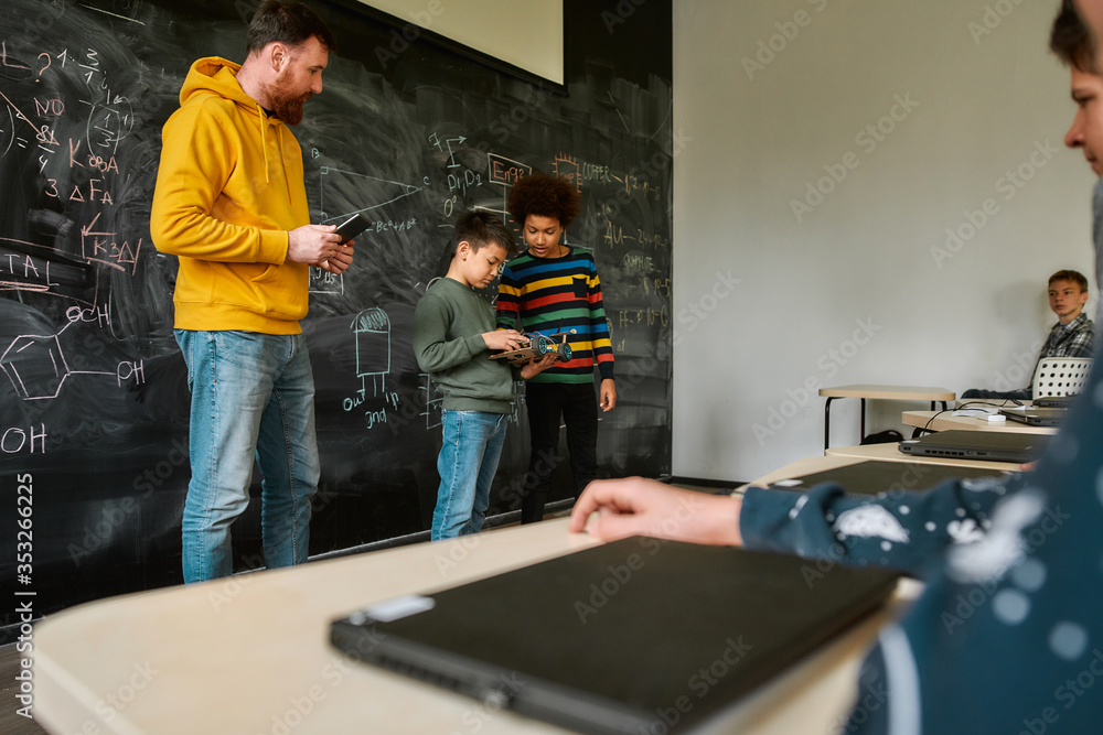 Center For Excellence. Young male science teacher standing near the blackboard, holding tablet pc and looking at his students while they are demonstrating their own robot vehicle