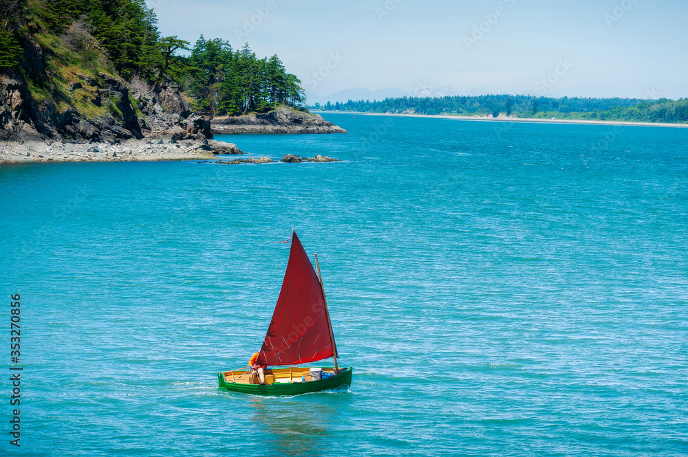 Obraz premium Sailboat on the Coast of Lummi Island, Washington. A sailboat departs Inati Bay on the east coast of Lummi Island heading towards Bellingham, Washington. Small boat with tanbark sail and wooden hull.