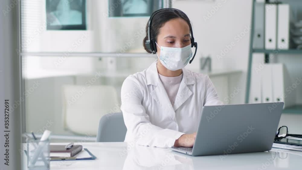Female physician in face mask, lab coat and wireless headset typing on laptop and talking via online call while working remotely with patient at desk in medical office Female physician in face mask, lab coat and wireless headset typing on laptop and talking via online call while working remotely with patient at desk in medical office
