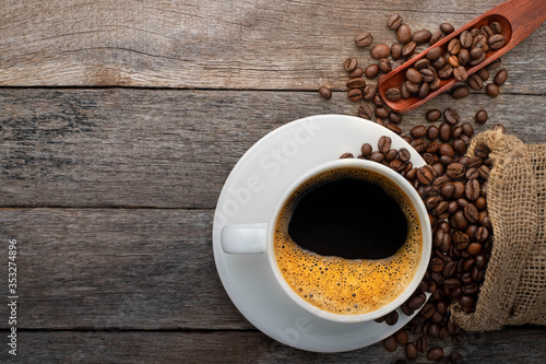 Top view of Hot fresh black coffee for morning with milk foam. Breakfast menu in white ceramic cup with coffee beans roasted in burlap sack bag on wood table background. Flat lay with copy space.