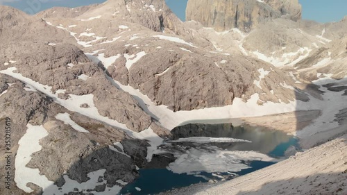 Aerial shot unveiling alpine lake Antermoia covered in ice from behind a ridge in Dolomites Italy. Aerial perspecitve of melting glacier in  South Tyrol.  photo