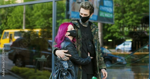 Cheerful Caucasian young stylish couple of hipsters walking in hungs at street during covid-19 pandemic. Beautiful extraordinary girlfriend with pink hair and boyfriend with tattoos strolling in masks photo