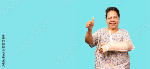 Elderly Asian woman broken arm cast  showing two thumbs up over blue background  in studio With copy space.
