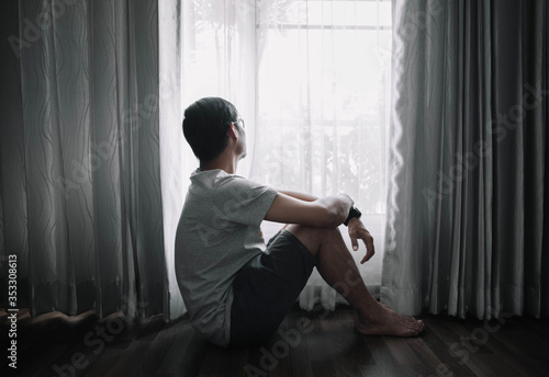 Sad and depressed young male sitting on the floor in the living room looking outside the doors,sad mood,feel tired, lonely and unhappy concept. Selective focus.
