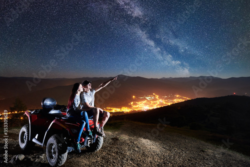 Romantic couple man and girl tourists sitting together on atv quad motorbike on the top of mountain. Man pointing at beautiful night sky full of stars, Milky way, luminous village on background