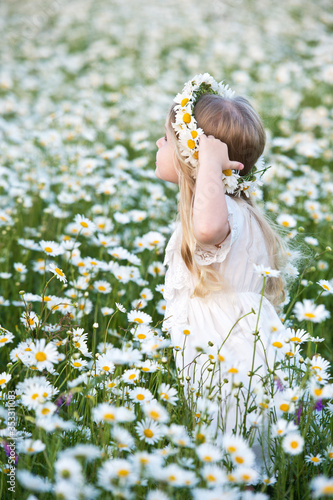 Beautiful girl with blond hairand chamomilie wreath in chamomile field. Portrait of cute little girl child with bouquet of chamomiles flowers in sunny summer day. photo