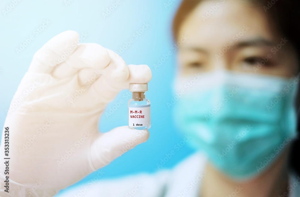A female Asian physician with surgical mask and white rubber gloves at a clinic, holding a glass bottle of 1 dose MMR vaccine with white background and red letters. A female Asian physician with surgical mask and white rubber gloves at a clinic, holding a glass bottle of 1 dose MMR vaccine with white background and red letters.