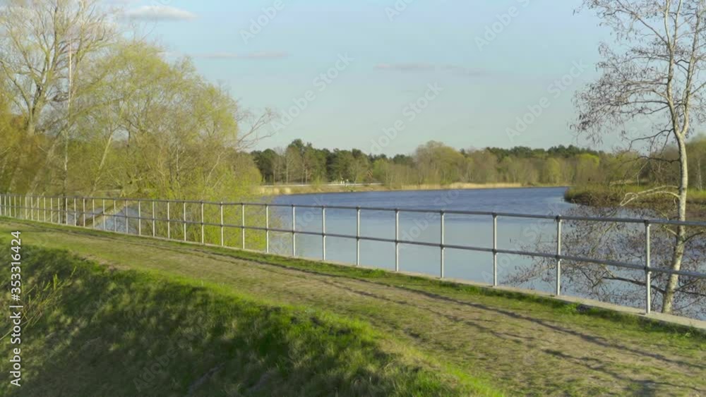 View Of Railing Alongside River Water Damn With Green Grass And Trees Against Blue Skies. Follow Shot View Of Railing Alongside River Water Damn With Green Grass And Trees Against Blue Skies. Follow Shot