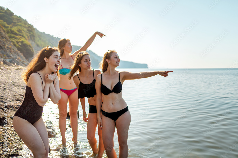 A group of beautiful young women in bathing suits look joyfully at the sea and point into the distance. In the background-the sea and sky. Copy space. Concept of vacation, holiday at the sea A group of beautiful young women in bathing suits look joyfully at the sea and point into the distance. In the background-the sea and sky. Copy space. Concept of vacation, holiday at the sea